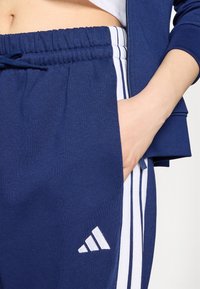 Tamsiai mėlynos sportinės kelnės su baltomis vertikaliomis juostelėmis, elastinga juosmeniu ir raišteliu. Kairiame šlaunyje yra baltas Adidas logotipas.