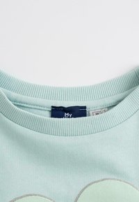 Sweatshirt vert menthe clair avec un col rond côtelé. Comprend une étiquette et une texture douce, avec des accents graphiques circulaires.