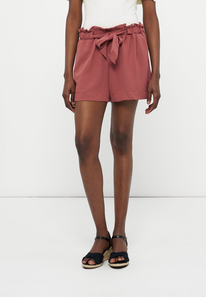 Γυναίκα που φοράει ψηλόμεσα σορτς σε απόχρωση σκόνης τριαντάφυλλου με ζώνη στη μέση, λευκή μπλούζα και μαύρα πλατφόρμες με λουράκι στον αστράγαλο, μπροστά σε λευκό φόντο.