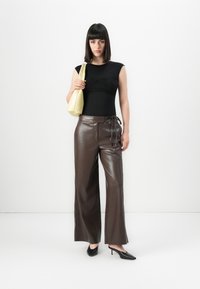 Donna con capelli lunghi e lisci di colore scuro che indossa un top senza maniche nero, pantaloni in pelle marroni a gamba larga, tacchi neri e porta una borsa a tracolla giallo chiaro.