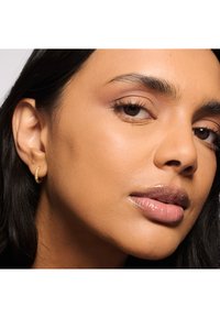 Pendiente de aro de oro con diseño torcido, llevado en una piel lisa de tono marrón claro. Primer plano del rostro, con un maquillaje natural.