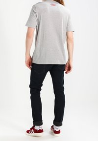 Camiseta de algodón gris de manga corta, con cuello redondo y un logo rojo en la parte posterior. Combinada con jeans oscuros y zapatillas rojas con detalles blancos.