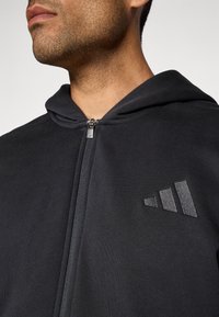 Homem vestido com uma hoodie preta com fecho de correr e um logo triangular bordado no lado esquerdo do peito, mostrado do pescoço até à metade do torso.