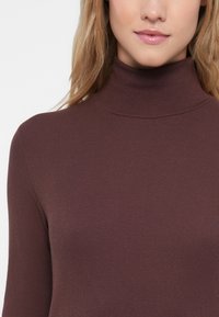 Dlhý rukáv rolák v tmavo burgundskej rebrovanej tkanine, s priliehavým strihom a jemnou textúrou. Detailný pohľad od ramien nahor.
