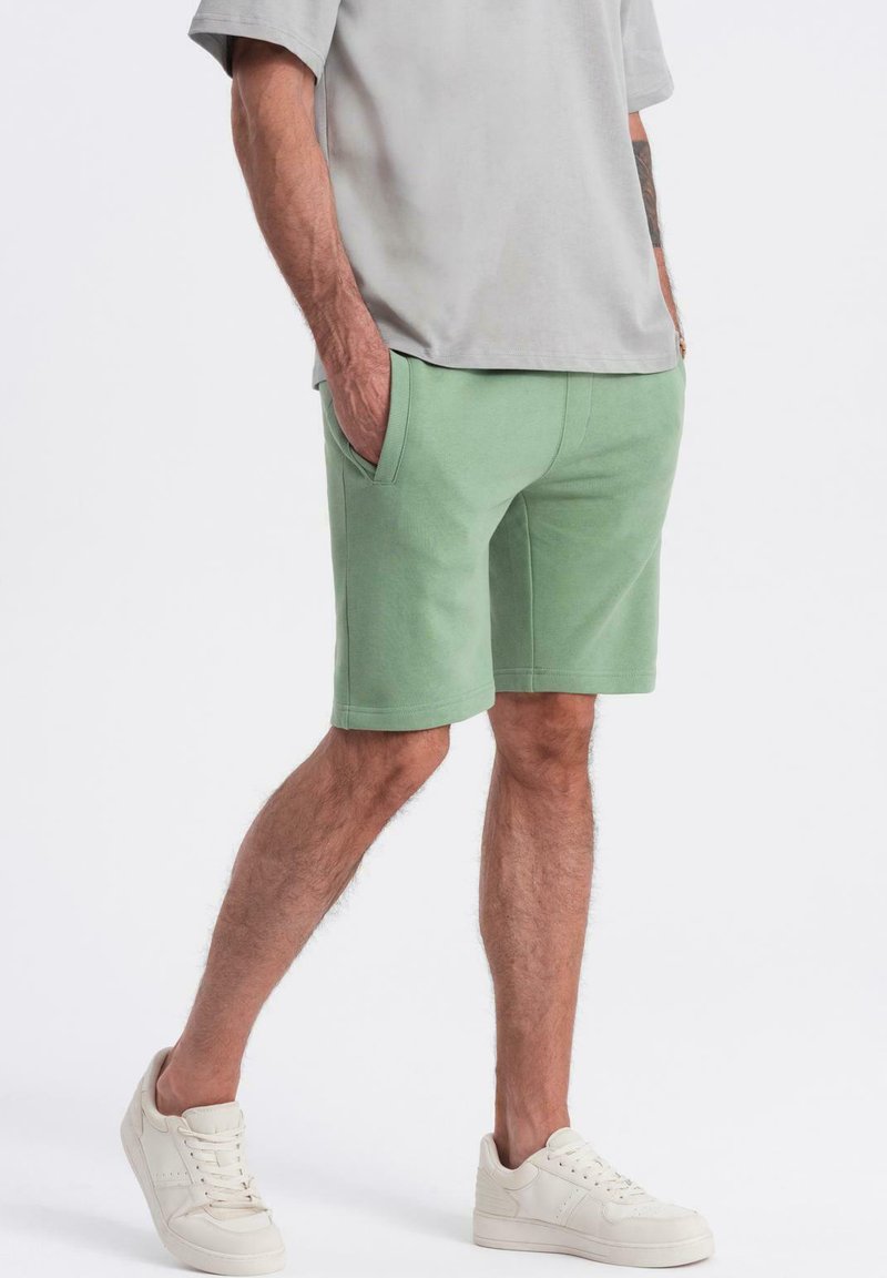 Shorts vert menthe en mélange de coton, avec une taille élastique, des poches latérales et une coupe décontractée. Associés à des baskets blanches.