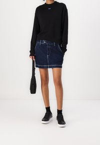 Schwarzes Sweatshirt, Jeans-Minirock, schwarze Sneakers und eine kleine schwarze Tasche mit Logo. Der Rock hat Gürtelschlaufen und kontrastierende Nähte.