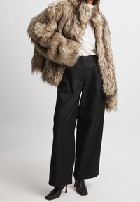 Bunda z umělého kožíšku v béžovo-hnědých tónech, s širokým límcem a nadýchanými rukávy, doplněná vysokými černými kalhotami a lodičkami na tenkém podpatku.