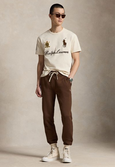 Νέος άνδρας που φοράει μπεζ μπλούζα Ralph Lauren, καφέ αθλητικό παντελόνι, λευκά ψηλά αθλητικά παπούτσια, γυαλιά ηλίου και ρολόι χειρός, στέκεται μπροστά σε απλό φόντο.