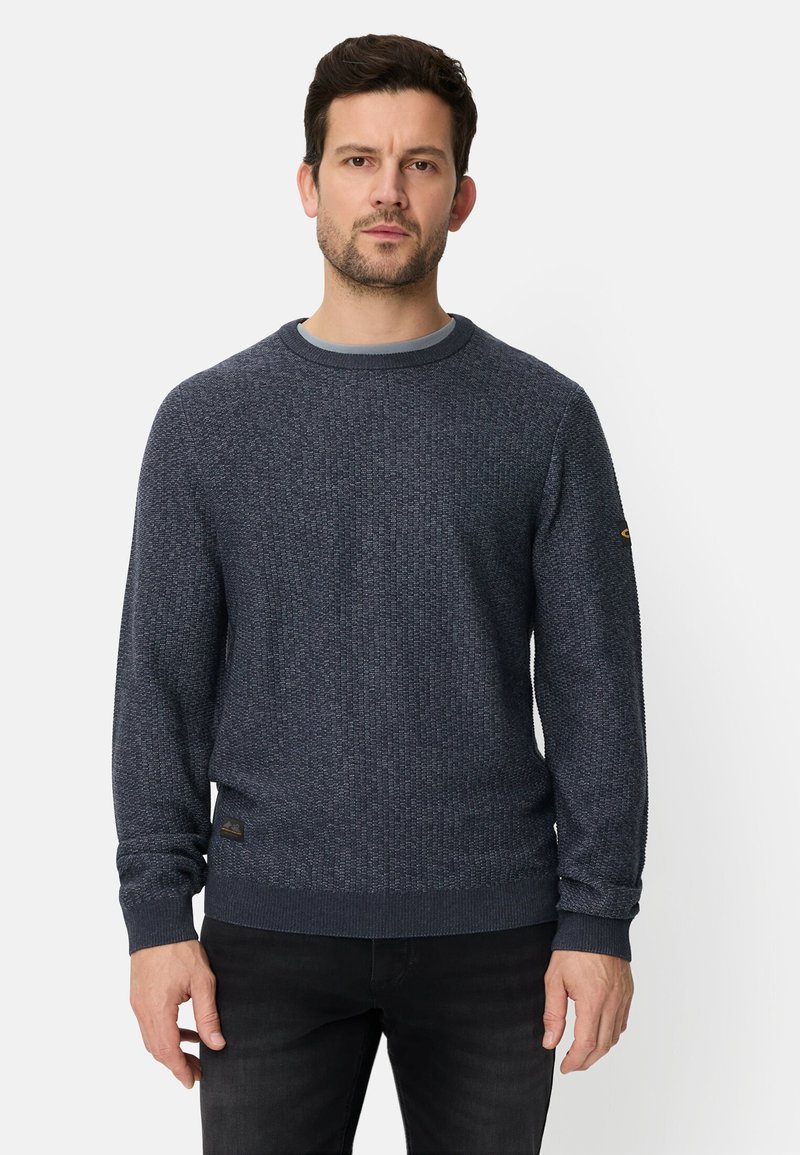 Granatowy sweter dziany z okrągłym dekoltem, teksturowaną tkaniną, ściągaczami na mankietach i dolnym brzegu oraz małym logo na rękawie. Noszony na jasnej koszuli.