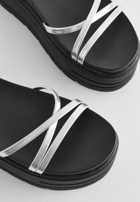 Juodos platformos sandalai su sidabro metalinėmis juostelėmis, kertančiomis pėdą. Tekstūruota padas su matoma sluoksnių konstrukcija.