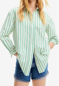 Camicia a righe verde e bianca con bottoni, realizzata in cotone, con colletto classico, maniche arrotolate e una vestibilità comoda. Indossata con pantaloni shorts in denim.