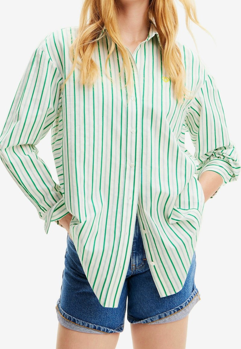 Camicia a righe verde e bianca con bottoni, realizzata in cotone, con colletto classico, maniche arrotolate e una vestibilità comoda. Indossata con pantaloni shorts in denim.