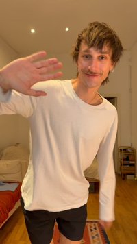 Jeune homme aux cheveux ébouriffés et à la moustache, agitant la main droite, portant une chemise blanche à manches longues et un short noir dans une pièce chaleureuse.