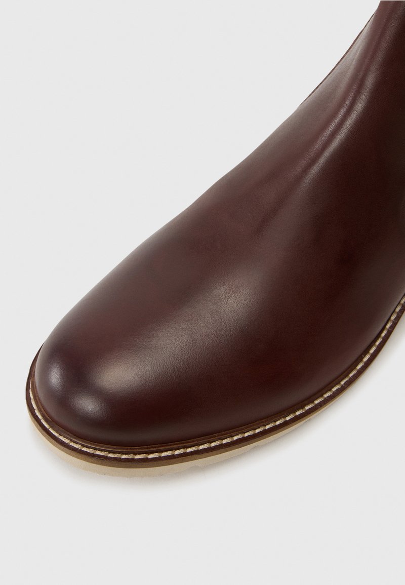Botte de cheville en cuir marron avec un bout arrondi, texture lisse et surpiqûres blanches contrastantes le long de la semelle, sur un fond clair.
