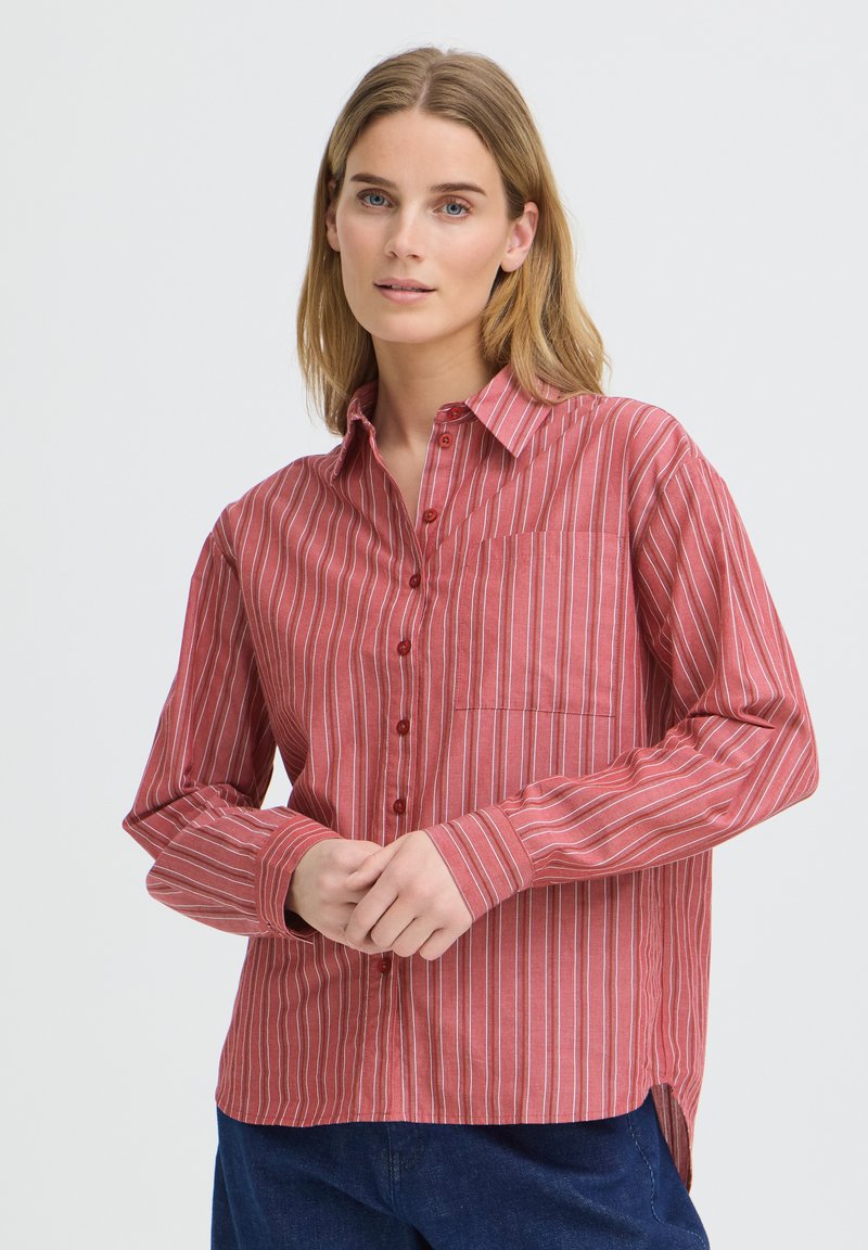 Femme aux cheveux châtain clair portant une chemise rouge à rayures blanches et un pantalon bleu foncé, debout les mains jointes devant elle.