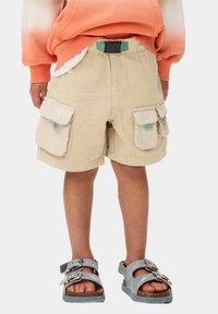 Beige cargo shorts med två sidofickor och ett svart justerbart bälte. Bärs med grå sandaler som har dubbla remmar och spännen.