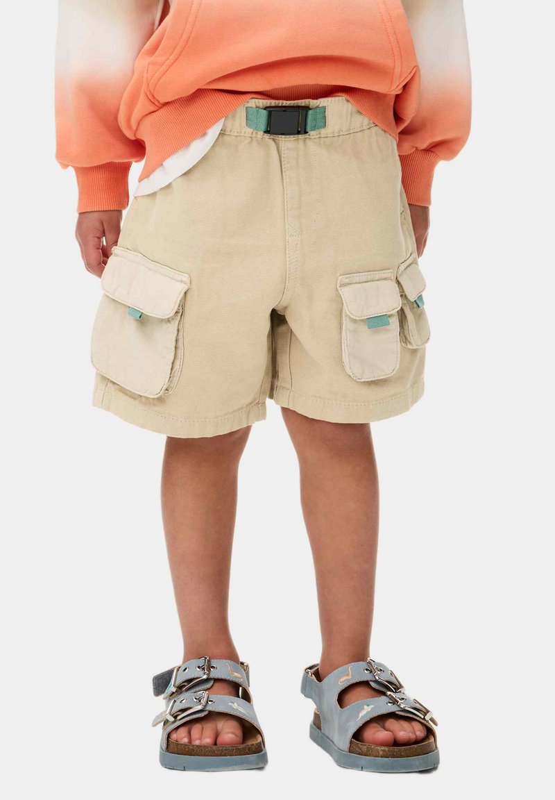 Beige cargo shorts med två sidofickor och ett svart justerbart bälte. Bärs med grå sandaler som har dubbla remmar och spännen.