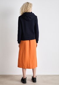 Femme aux cheveux blonds bouclés portant un sweat à capuche noir, une jupe midi orange et des baskets noires, debout face à un mur blanc.