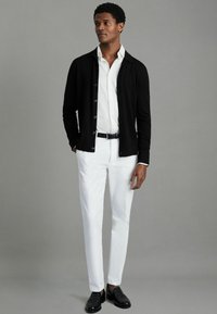 Cardigan negru cu mâneci lungi peste o cămașă albă, asociat cu pantaloni albi și pantofi negri. Texturi netede și design ajustat.