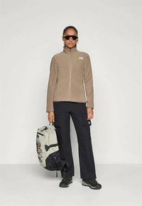 Person in beigem Reißverschlussjacke, schwarzen Cargohosen, schwarzen Wanderschuhen und großen Sonnenbrillen, hält einen hellgrauen und schwarzen Rucksack an der Seite.