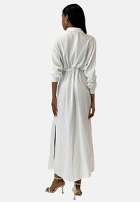 Vestido largo de algodón blanco con un acabado texturizado, cuello con botones, cinturilla con lazo y aberturas laterales, combinado con sandalias de tacón alto en tono nude.