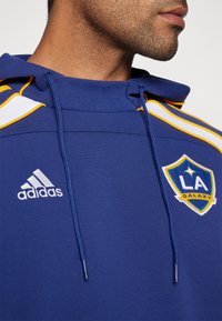 Blue hoodie made of cotton blend, featuring white and yellow stripes on shoulders, Adidas logo, and LA Galaxy emblem embroidered prominently.