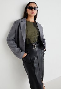Une veste oversize grise sur un top vert olive, associée à une jupe en cuir noir avec une fente à l'avant et une ceinture à boucle dorée. Lunettes de soleil noires.
