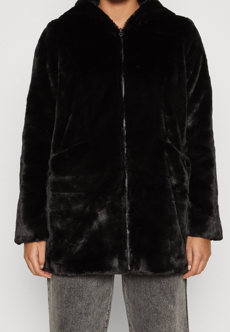 Personne portant une veste en peluche noire à fermeture éclair avec poches à l'avant et manches longues, associée à un jean gris délavé.