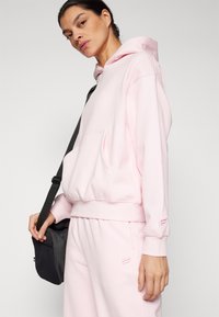 Felpa rosa realizzata in tessuto morbido, con tasca frontale e polsini a costine, abbinata a pantaloni della tuta coordinati e una borsa a tracolla nera.