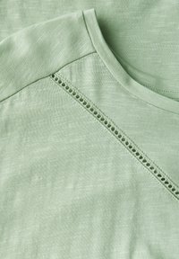 Světle zelené tričko s jemnou texturou, kulatým výstřihem a dekorativními otvory podél švu na rameni.
