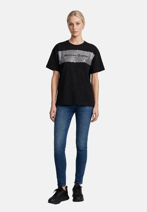 Mujer con cabello rubio que lleva una camiseta negra de Maison Gaelle Paris, vaqueros pitillo azules y zapatillas negras, posando frente a un fondo liso.