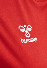 Röd sporttröja i tyg med en vit Hummel-logotyp, som visar en geometrisk design med stiliserade vingar och chevrar. Mjuk, texturerad yta.