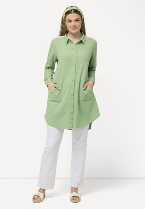 Lichtgroene button-up tuniek met lange mouwen, twee voorzakken en een afgeronde zoom, gepaard met witte broek en sandalen.