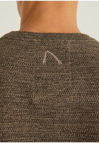 Gros plan sur un pull en maille texturée marron montrant l'encolure arrière et un petit logo triangulaire brodé de couleur claire.