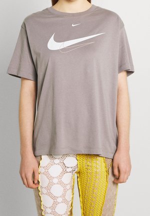 Osoba nosząca szarą koszulkę Nike z warstwowymi logo Swoosh oraz żółte i białe spodnie z szydełkowym patchworkiem, stojąca na jednolitym tle.