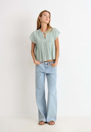 Vrouw met lang haar die een lichtgroen geborduurd blouse met korte mouwen, lichtblauwe wijde jeans, sandalen en gouden oorbellen draagt, staand.