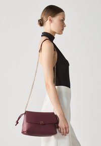 Sac à main en cuir bordeaux avec une sangle nouée, une chaîne dorée et un fermoir à rabat. Texture lisse, forme rectangulaire et design minimaliste.