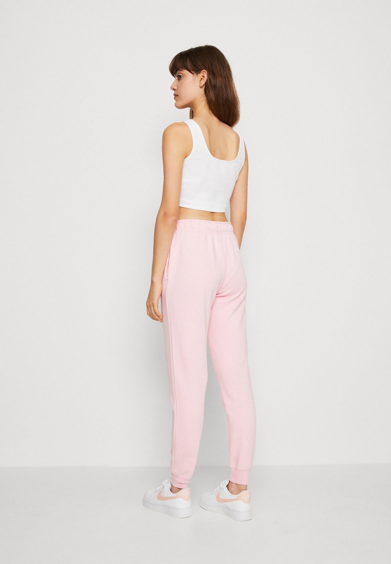 Femme se tenant de côté, portant un crop top blanc, un pantalon de jogging rose clair et des baskets blanches avec des accents rose clair, sur un fond uni.