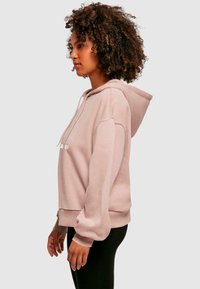 Rózsaszín kapucnis pulóver, zsinórral, túlméretezett fazon, bordás mandzsettákkal. Fekete leggings-szel viselve, amely laza dizájnt és puha textúrát mutat.