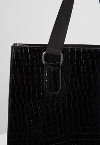 Sac à main noir avec une texture crocodile brillante et une large bandoulière en tissu noir attachée par un anneau métallique rectangulaire argenté.