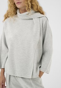 Világosszürke, textúrás pulóver dropt nyakkal és széles ujjakkal. Oldalsó panel részletek és aszimmetrikus alsó él jellemzi.