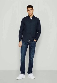Mørkeblå knap-opsat skjorte, lange ærmer, let stof; sat sammen med mørke denim jeans og hvide sneakers, stående pose, neutral baggrund.