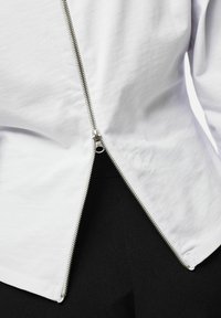 Chemise en coton blanc avec une fermeture éclair argentée sur le côté, présentant un ourlet arrondi et une texture lisse. Un tissu noir est visible en dessous.