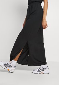 Pantalon noir à jambes larges avec fentes latérales à l'ourlet, associé à des baskets blanches avec des accents noirs et orange.