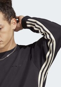 Svart sweatshirt med krämfärgade randade detaljer längs ärmarna. Mjuk textur med rund hals och minimal logotyp på bröstet.