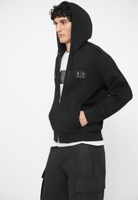 Mlad muž nosi crnu duksericu sa zatvaračem i kapuljačom, ispod bijelu majicu, i crne cargo hlače, stoji s jednom rukom u džepu.