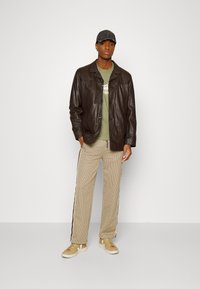 Bruine leren jas over een olijfgroen t-shirt, beige geruite broek met zijstrepen, en beige sneakers met witte zolen.