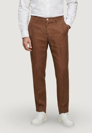 Uomo con pantaloni marroni su misura, camicia bianca con bottoni e sneaker bianche, con una mano in tasca, in piedi davanti a uno sfondo semplice.