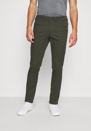 Chinos verts olive pour hommes en coton. Coupe slim avec une fermeture éclair, un bouton et des passants de ceinture. Associés à des baskets blanches.
