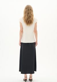 Haut blanc à manches courtes associé à une longue jupe noire. La jupe a une texture lisse, et la tenue est complétée par des talons noirs.